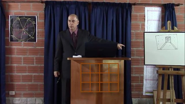 Un hombre calvo vestido con traje oscuro y corbata granate, de pie frente a un podio de madera en una sala con cortinas azules. A su izquierda, hay un cuadro de la Mazzaroth y a su derecha, un caballete con un dibujo de un matraz Erlenmeyer y una estructura atómica. Öffnet ein Video in einem neuen Fenster.