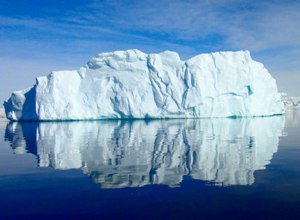 Un gran iceberg flotante sobre aguas tranquilas bajo un cielo despejado, reflejando una simetr&iacute;a perfecta entre el cielo y el mar.