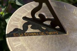 Reloj de sol de metal con la frase "En la sombra del tiempo" grabada, mostrando la sombra proyectada por el gnomon que indica la hora solar. Interner Link zu einem Artikel auf dieser Website.
