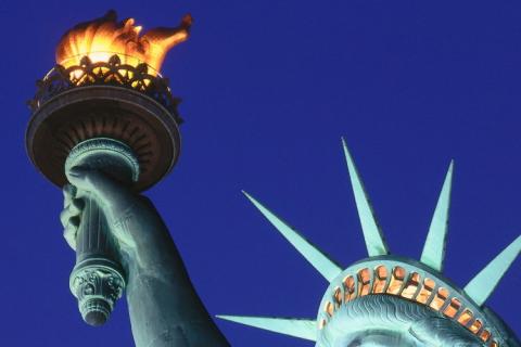 Vista cercana de la antorcha encendida y la corona de la Estatua de la Libertad contra un cielo azul nocturno, simbolizando la iluminaci&oacute;n y la libertad bajo las estrellas.