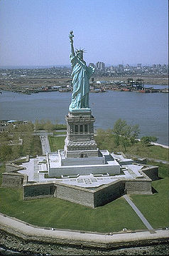 Vista a&eacute;rea de la Estatua de la Libertad, un gran monumento de una figura femenina con una antorcha elevada, ubicada en una isla en el centro de un cuerpo de agua, con una ciudad visible al fondo.