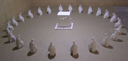 Veinticuatro estatuas peque&ntilde;as con capas blancas dispuestas en un patr&oacute;n circular alrededor de un cubo blanco iluminado en el centro, sobre un suelo de tierra en un espacio cerrado con paredes de concreto.