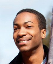 Joven afroamericano sonriendo a la c&aacute;mara, con luz solar suave iluminando su rostro y un fondo desenfocado claro.