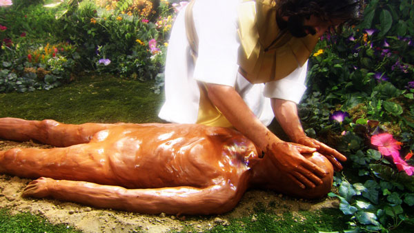 Imagen de una representaci&oacute;n art&iacute;stica donde una persona con t&uacute;nica blanca y larga cabellera est&aacute; tocando a una figura humana tendida sobre el suelo en un jard&iacute;n florecido. La escena transmite un sentido de curaci&oacute;n o cuidado.