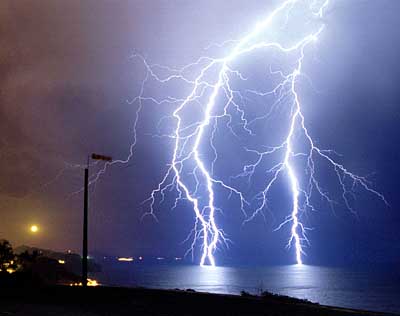 Rel&aacute;mpagos intensos iluminando el cielo nocturno sobre el mar, con luces tenues al fondo en una costa.