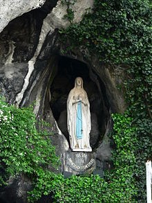 Estatua de una figura religiosa femenina colocada en una gruta artificial rodeada de plantas verdes. Sobre la figura se puede ver una leyenda que se refiere al Mazzaroth en tonos azul y blanco.