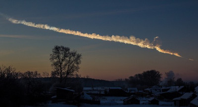 Una estela brillante en el cielo crepuscular sobre un paisaje invernal con &aacute;rboles desnudos y casas cubiertas de nieve, sugiriendo un fen&oacute;meno celestial o un cuerpo celeste en movimiento.