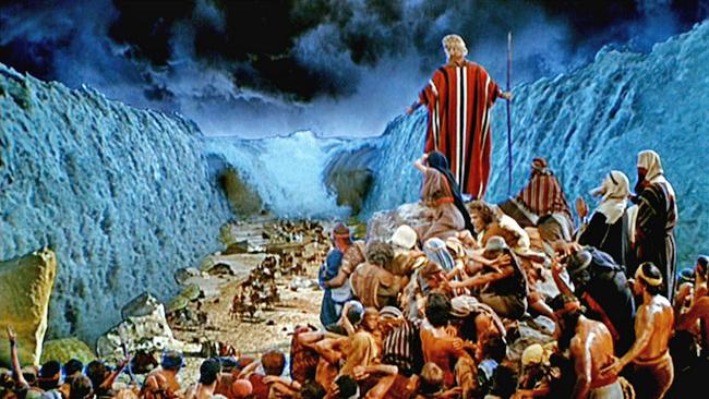 Imagen cinematogr&aacute;fica de una escena b&iacute;blica donde un l&iacute;der, vestido con t&uacute;nicas rayadas en rojo, blanco y azul, se alza ante una multitud de personas que atraviesan entre dos enormes muros de agua que forman un ca&ntilde;&oacute;n. El cielo est&aacute; nublado y oscuro, agregando un tono dram&aacute;tico a la escena.