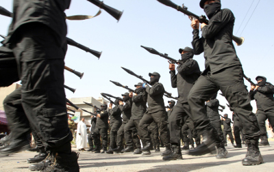 Un grupo de hombres vestidos con uniformes negros marchando en formaci&oacute;n mientras sostienen rifles hacia arriba, en una calle pavimentada durante un d&iacute;a soleado.