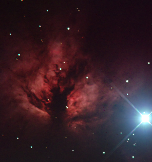 Imagen de una nebulosa brillante y compleja, te&ntilde;ida en tonos de rojo y negro con estrellas dispersas alrededor, y un brillante rayo de luz blanca en la esquina inferior derecha.