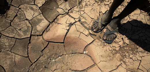 Suelo &aacute;rido y cuarteado visto desde arriba, con dos pies descalzos de una persona parados sobre la tierra seca. Los pies tienen sandalias simples.