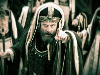 Hombre con vestimenta tradicional jud&iacute;a, incluyendo un tocado decorado, se&ntilde;alando hacia adelante con expresi&oacute;n intensa en un entorno de reuni&oacute;n. La imagen tiene un matiz sepia que a&ntilde;ade un efecto antiguo.