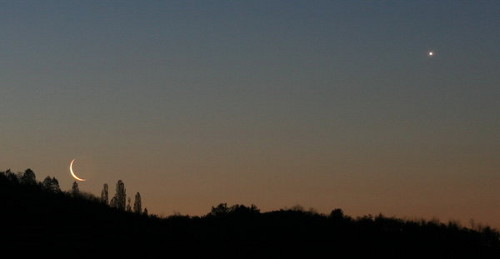 Imagen de un crep&uacute;sculo con una luna creciente muy delgada y un brillante planeta visible en el cielo sobre un horizonte oscuro con siluetas de &aacute;rboles.