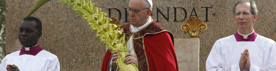 Tres cl&eacute;rigos en una ceremonia religiosa al aire libre, uno de ellos sostiene una gran rama de palma, vestidos con ropas lit&uacute;rgicas rojas y moradas, frente a un fondo de piedra con texto grabado. Interner Link zu einem Artikel auf dieser Website.