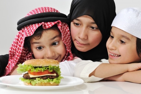 Tres ni&ntilde;os de diferentes edades, vestidos con ropas tradicionales, observan con inter&eacute;s una hamburguesa en un plato blanco sobre una mesa. El ambiente es luminoso y alegre.
