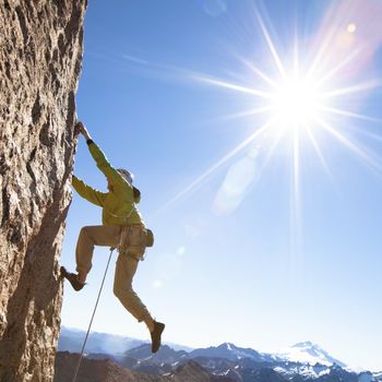 Un escalador ascendiendo una pared rocosa, vestido con equipo para escalar, bajo un sol brillante. Monta&ntilde;as nevadas se ven en la distancia bajo un cielo despejado.
