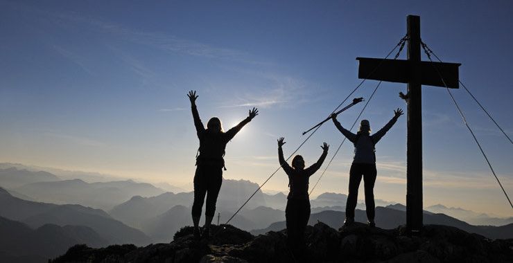 Tres personas celebran levantando las manos en la cima de una monta&ntilde;a al amanecer, con una cruz de madera en el centro y las monta&ntilde;as distantes en el fondo bajo un cielo amplio.