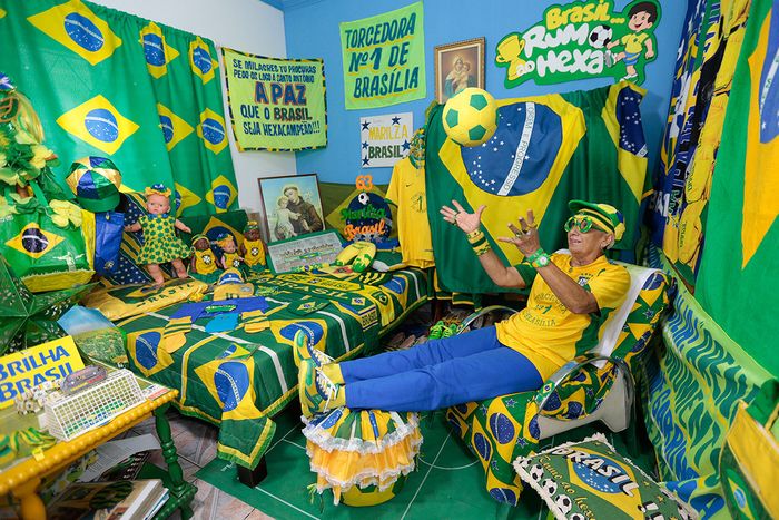 Una persona emocionada viste un atuendo tem&aacute;tico de f&uacute;tbol en colores verde y amarillo, dentro de una habitaci&oacute;n decorada profusamente con banderas, mantas y objetos similares alusivos a Brasil. La habitaci&oacute;n resalta por su vibrante paleta de colores y diversos elementos con el tema del f&uacute;tbol.