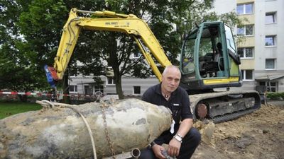 Un hombre calvo agachado al lado de un antiguo proyectil sin detonar, con una excavadora amarilla al fondo, situada en una zona de construcci&oacute;n urbana.