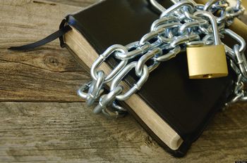 Un libro cerrado con tapa negra est&aacute; asegurado con una cadena de eslabones de metal y un candado dorado sobre una superficie de madera r&uacute;stica.