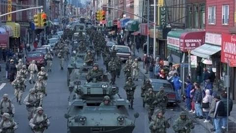 Convoy militar con varias tanquetas y tropas armadas transitando por una calle urbana con tiendas y peatones observando.