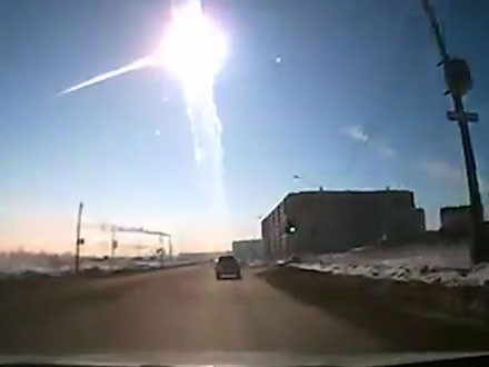 Imagen capturada desde el interior de un veh&iacute;culo que muestra una carretera con nieve a los lados, un edificio industrial y un cielo claro con un brillante meteoro descendiendo diagonalmente en un fen&oacute;meno celeste.
