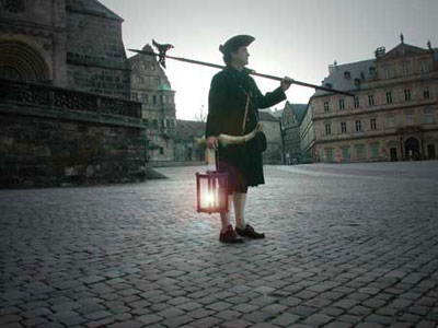 Un hombre vestido con indumentaria hist&oacute;rica sostiene una l&aacute;mpara encendida en una calle adoquinada, bajo un cielo crepuscular que podr&iacute;a sugerir el misterio de Mazzaroth.