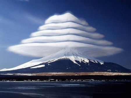 Imagen de un volc&aacute;n cubierto de nieve con m&uacute;ltiples nubes lenticulares apiladas verticalmente encima, creando un efecto visual escalonado sobre un cielo azul.