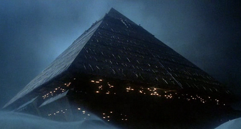 Una imagen de una gran estructura piramidal iluminada por m&uacute;ltiples luces interiores, emergiendo de una neblina densa bajo un cielo oscuro.