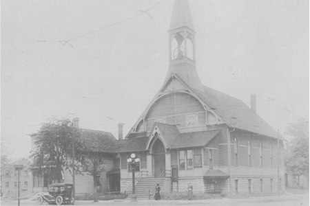 Fotograf&iacute;a en blanco y negro de una iglesia de madera con un steeple prominente y un coche antiguo estacionado al frente. Varias personas est&aacute;n paradas cerca de la entrada.