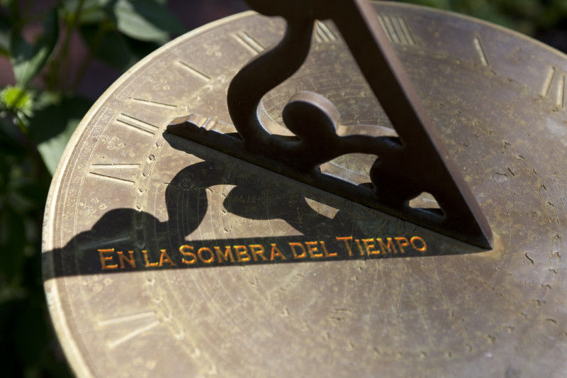 Una imagen de un reloj solar con una figura estilizada que proyecta una sombra sobre una plancha met&aacute;lica inscrita con n&uacute;meros romanos y elementos de la Mazzaroth. Una frase en la superficie dice: "En la sombra del tiempo".
