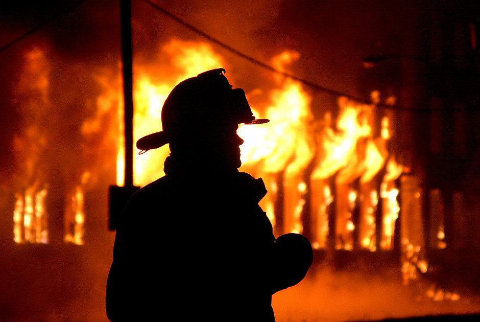 Silueta de un bombero de pie frente a un incendio intenso en un edificio durante la noche, con llamas brillantes iluminando el fondo oscuro.