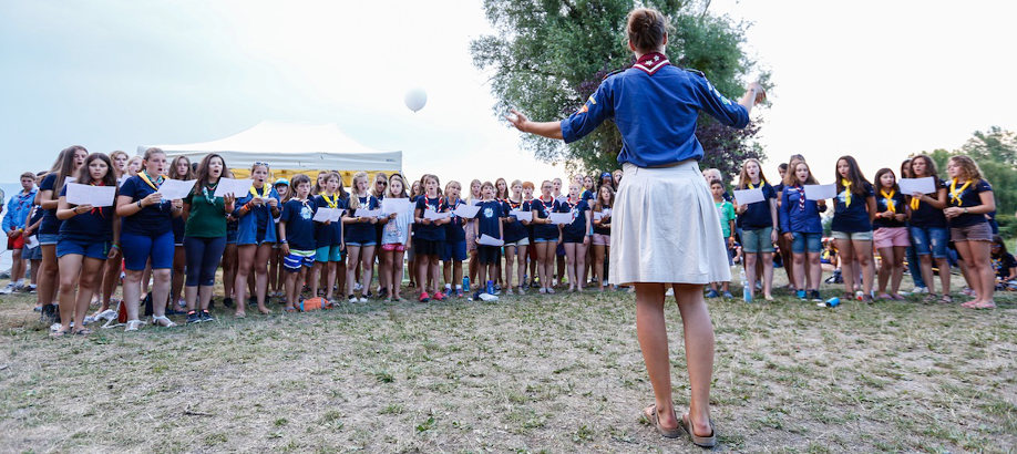 Una mujer de pie, de espaldas, dirige a un grupo de j&oacute;venes en uniformes azules agrupados al aire libre, algunos sosteniendo papeles. &Aacute;rboles verdes y cielo despejado en el fondo.