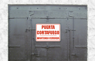 Puerta met&aacute;lica industrial de color gris oscuro con un cartel rojo que dice "PUERTA CORTAFUEGO MANTENGA CERRADA" ubicada en una pared de azulejos blancos.