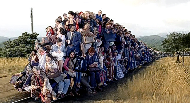 Imagen de una larga fila de personas sentadas una tras otra en el suelo de un camino rural, extendi&eacute;ndose hacia el horizonte entre colinas suaves. Las personas muestran diversas expresiones y estilos de vestimenta, sugiriendo una convocatoria diversa y numerosa bajo el cielo claro.