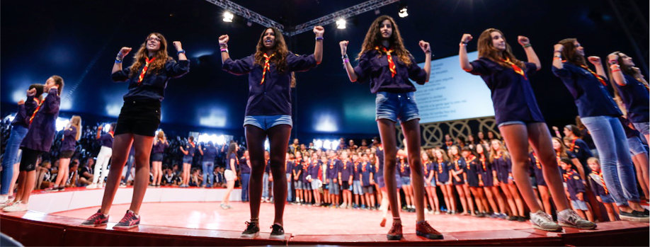 Grupo de j&oacute;venes estudiantes emocionados, levantando los pu&ntilde;os en el aire mientras est&aacute;n en un escenario iluminado, con un grupo grande de personas en el fondo dentro de un auditorio.