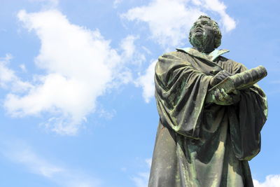 Estatua de bronce de un hombre con barba y toga, mirando hacia el cielo, con las manos cruzadas sobre un rollo, contra un fondo de cielo azul con nubes dispersas.