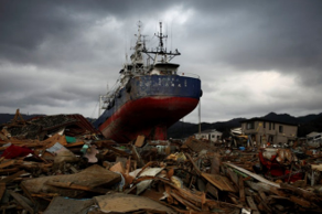 Un barco de gran tama&ntilde;o con un casco rojo y azul encallado en medio de escombros de madera y estructuras destruidas bajo un cielo nublado que evoca la turbulencia celestial.