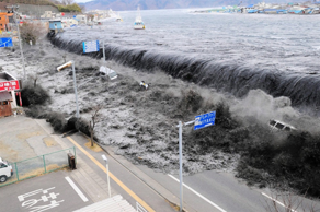 Vista a&eacute;rea de un enorme tsunami avanzando hacia una costa urbana con edificios y se&ntilde;ales de tr&aacute;fico visibles, demostrando la poderosa fuerza de la naturaleza.
