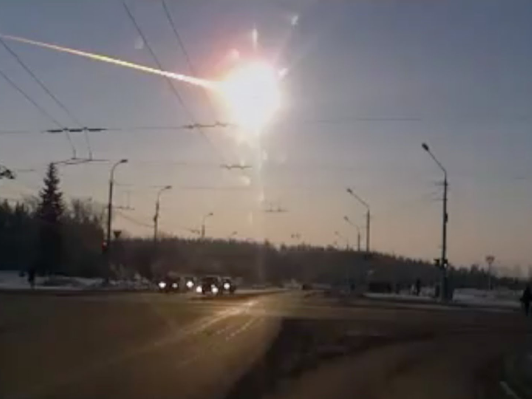Una vista de una calle concurrida durante el atardecer, mostrando el sol brillante con un rastro luminoso en el cielo, posiblemente un meteorito, mientras los veh&iacute;culos circulan y las farolas alineadas a lo largo de la v&iacute;a est&aacute;n encendidas.