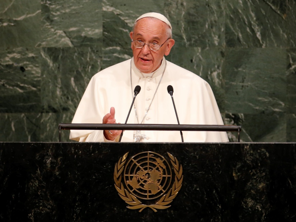 Un hombre vestido de blanco habla desde un podio con el emblema de las Naciones Unidas en un fondo de m&aacute;rmol verde oscuro.
