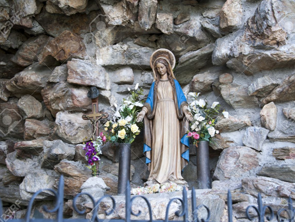 Una estatua de Mar&iacute;a, con halo y vestida con ropas de tonos claros y azules, est&aacute; rodeada de flores multicolores y se ubica frente a un fondo de rocas irregulares. Un peque&ntilde;o crucifijo de metal acompa&ntilde;a la escena situado a la izquierda de la figura.