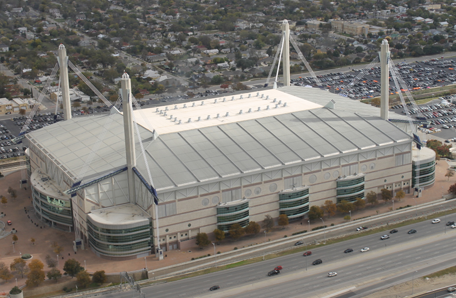 Vista a&eacute;rea de un gran estadio con techo blanco y estructura de soporte tipo torre, rodeado de estacionamientos abarrotados y v&iacute;as de tr&aacute;fico activas.