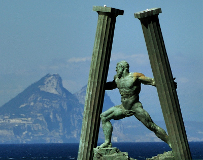 Escultura de bronce de un hombre con barba en pose din&aacute;mica, ubicada entre dos columnas de piedra desgastadas con un fondo de monta&ntilde;a y mar azul.