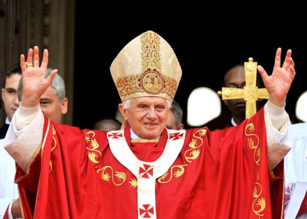 Un hombre de edad avanzada vestido con ornamentos lit&uacute;rgicos rojos y una mitra dorada, levanta las manos en un gesto de bendici&oacute;n frente a una multitud.