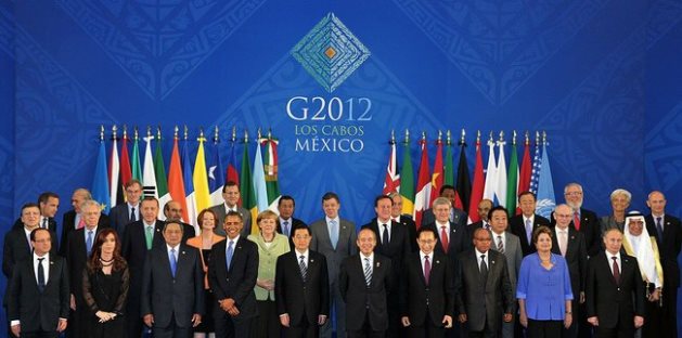 Foto oficial del evento G20 2012 en Los Cabos, M&eacute;xico, mostrando a l&iacute;deres mundiales de pie en varias filas frente a banderas internacionales y el logotipo del evento sobre un fondo azul decorado.