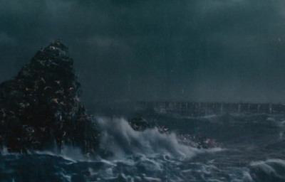 Una imagen de una tumultuosa escena oce&aacute;nica durante una tormenta, con grandes olas chocando contra una roca en primer plano y un puente largo y oscuro al fondo bajo un cielo cubierto.