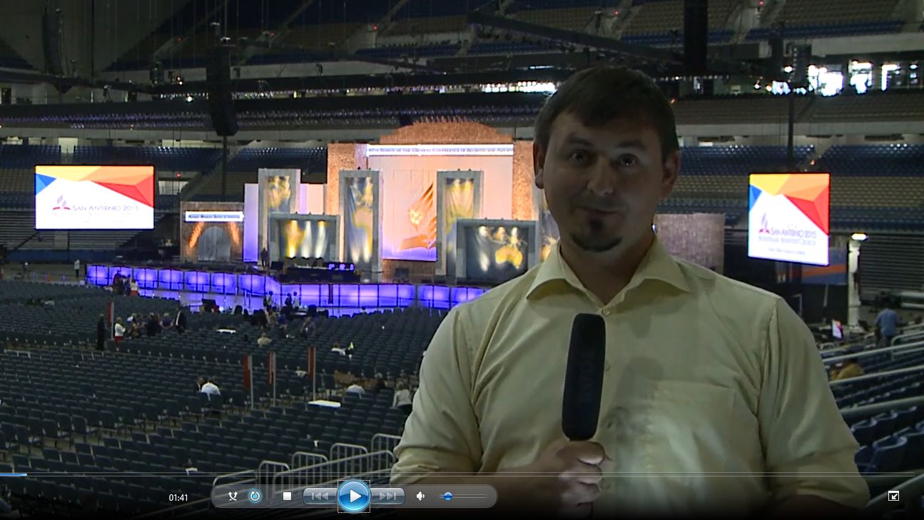 Un hombre con camisa amarilla est&aacute; de pie sosteniendo un micr&oacute;fono en primer plano, con un escenario grande en el fondo dentro de un estadio. El escenario est&aacute; iluminado con im&aacute;genes de Mazzaroth y banners promocionales del evento.
