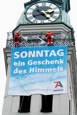 Foto de un reloj de torre grande con la hora apuntando hacia las diez y dieciocho. Dos figuras vestidas de Santa Claus est&aacute;n decorando la estructura. Debajo del reloj, cuelga un cartel grande en alem&aacute;n que dice "SONNTAG ein Geschenk des Himmels", promocionando la observaci&oacute;n del domingo.