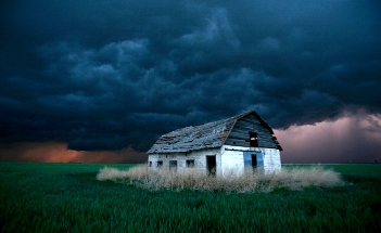 Una casa desolada y abandonada con tejado a dos aguas se encuentra en un campo de hierba bajo un cielo oscuro y amenazador, simbolizando un momento de tranquilidad antes de la tormenta.